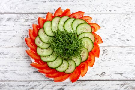 Beautiful sliced vegetables for the festive table. Sliced red tomatoes, green cucumbers, dill. Plate with vegetables on a white wooden background.の写真素材