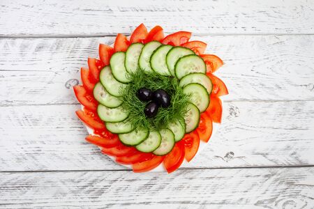 Beautiful sliced vegetables for the festive table. Sliced tomatoes, cucumbers, dill, olives. Plate with vegetables on a white wooden background.の写真素材