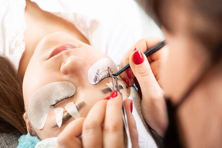 Eyelash extensions in a beauty salon. A young girl lies on a couch with her eyes closed. The master glues bundles of eyelashes using black glue.の写真素材