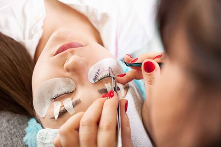 Eyelash extensions in a beauty salon. A young girl lies on a couch with her eyes closed. The master glues bundles of eyelashes using black glue.の写真素材