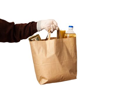 A volunteer delivers food to those in need. Delivery donation. Sunflower oil, canned food, pasta in an eco-friendly paper bag. Hands in gloves. Isolate on a white background.の写真素材