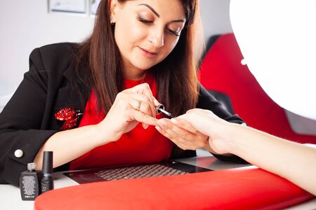 The woman covers the client's nails with a transparent varnish fixer. The process of manicure in a beauty salon. The master of manicure puts in order the nails of the client.の写真素材