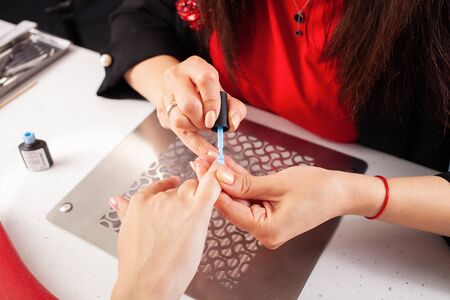 The woman covers the client's nails with blue gel varnish. The process of manicure in a beauty salon. Salon in black, red, white. The master of manicure puts in order the nails of the client.の写真素材