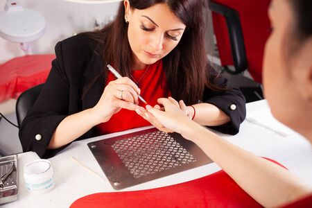 The woman lubricates the client's nails with a brush. The process of manicure in a beauty salon. Salon in black, red, white. The master of manicure puts in order the nails of the client.の写真素材