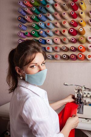 The seamstress in the mask overlays the edge of the fabric of the future red dress. A woman in a sewing studio sits behind an overlock. Small businesses in the face of the coronavirus pandemic.の写真素材