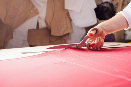 A seamstress woman cuts off the excess red fabric, the girl in the atelier cuts out the details of...