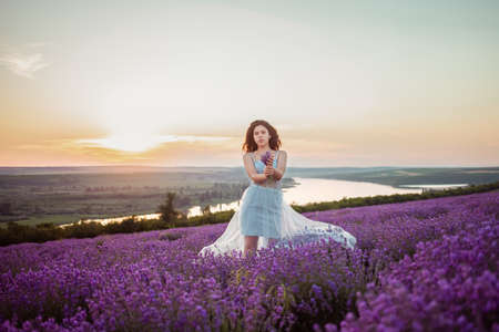 A beautiful young girl against the sunset and a beautiful sky in a lavender field. Purple lavender, orange sky, empty space for text. Advertising photo of perfume, makeup, fashion, lavender, beauty.の写真素材