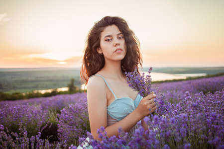 A beautiful young girl against the sunset and a beautiful sky in a lavender field. Purple lavender, orange sky, empty space for text. Advertising photo of perfume, makeup, fashion, lavender, beauty.の写真素材
