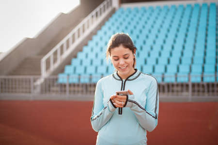 Beautiful girl looks in the phone on the background of the stadium in the sunset. The girl in a blue sports uniform smiles after a workout.の写真素材