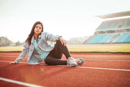 Beautiful girl looks in the phone on the background of the stadium in the sunset. The girl in a blue...