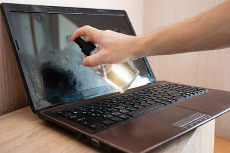 The man cleans the laptop from dust and bacteria with alcohol spray. Disinfection, protection...