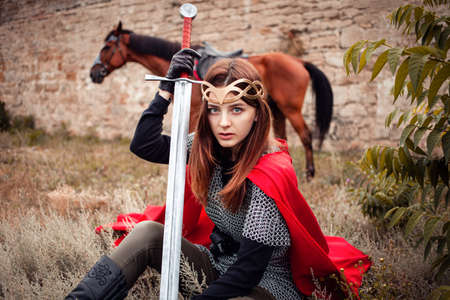 A girl in a medieval suit looks into the camera, sits on the grass, a horse grazes from behind in the background of the fortress. A woman with a sword in her hand, in a chain mailの写真素材