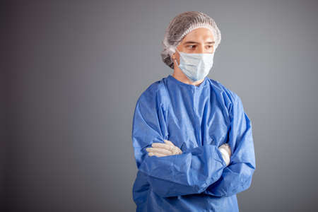 A handsome male surgeon is a doctor on a gray background in the studio. A man in a robe, a medical mask, a hat, gloves. Empty space for text. A man stands with his arms folded on his chest.の写真素材