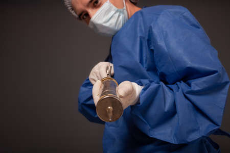 A male doctor with a terrible old syringe in his hands. The doctor in a medical mask, hat, robe and gloves. A terrible. The doctor scares with a huge syringe with a thick needle.の写真素材