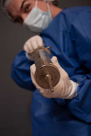 A male doctor with a terrible old syringe in his hands. The doctor in a medical mask, hat, robe and...