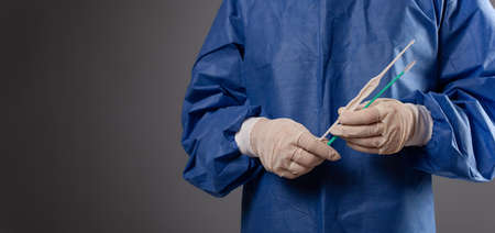 A gynecologist with a venus mirror in his hands. Gynecological accessories, brush, stick. A doctor in a medical mask, a robe, gloves, with a gynecological mirror on a gray background.の写真素材