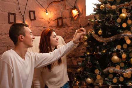 Young people in love hang a toy on the Christmas tree. A guy and a girl dress up a Christmas tree for Christmas. New Year's decorations in the photo studio. Posing for models.の写真素材