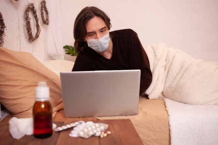 A sick man in a medical mask against the background of tablets. Home quarantine, coronavirus, covid. A man lies on a bed with a laptop.の写真素材