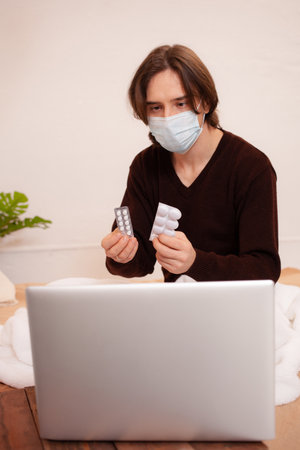 A sick man sits in front of a laptop and shows the doctor his pills. Home quarantine, medical online consultation. A man wearing a mask at home.の写真素材