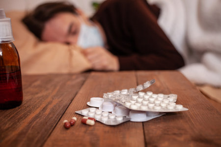 A sick man in a mask against a background of pills and medicines. The man is being treated at home in quarantine. Coronavirus, covid, cold, flu. A man fell ill in a home quarantine.の写真素材