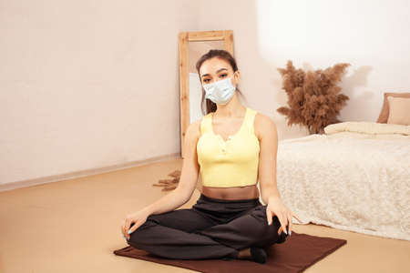 Young beautiful sports girl in beige interior. Yellow gray clothing. A healthy lifestyle. A woman in a medical mask is engaged in yoga fitness.の写真素材