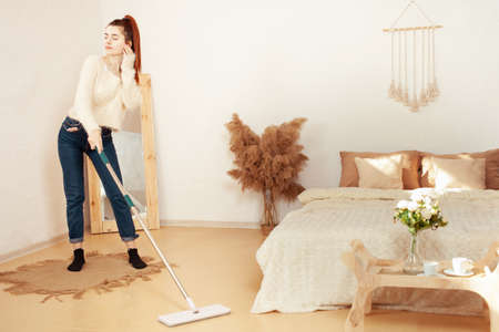 A beautiful young woman cleans the house and listens to music. Beige interior, funny girl. A woman washes the floor with a mop.の写真素材