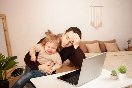 A man works at home on a laptop. The child shouts yells hysterically prevents distractions from work. The father can not cope with his daughter, angry, tired. Freelance on a computer interiorの写真素材