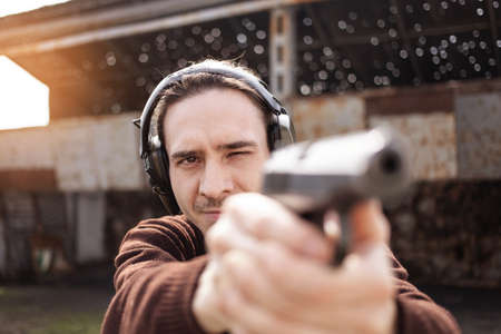 A young man shoots a gun, aiming at the target. A man wearing protective headphones. A wall and a roof with bullet holes. outdoor shooting range close upの写真素材
