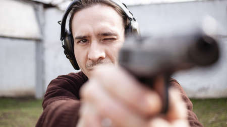 A young man is aiming for a gun. A man wearing protective headphones. outdoor tire shooting range. hobby. firearm against the white wall close upの写真素材