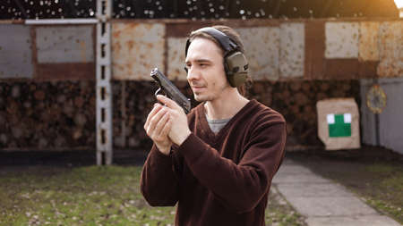 A young man shoots a gun, aiming at the target. A man wearing protective headphones. A wall and a...