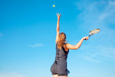 A beautiful young woman plays tennis on the court. Sporting a black suit dress. Blue sky, empty space for text. The girl is giving the ball. Exercise, active lifestyle, concept. portrait. mocupの写真素材
