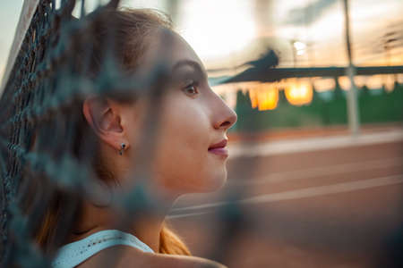 Portrait of a beautiful girl. Athlete on the tennis court. Grid, sunset. rest after training. Smiles, happy, sunny glare. Active lifestyle, outdoor. mesh in the foregroundの写真素材