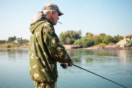 The older man is fishing on the river. Dirty water, pollution, garbage, rusty barges. Ecological catastrophe, production waste. senior concept, smokesの写真素材