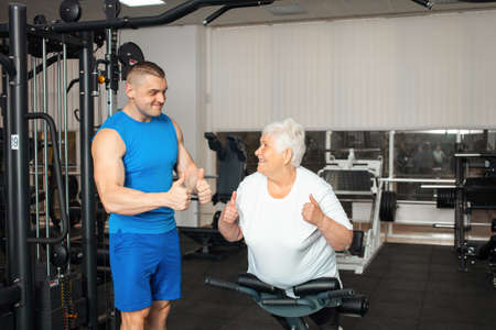 An elderly woman rocks the abs on the simulator in the gym. Happy smiling people. Healthy active sports lifestyle, senior concept. Rehabilitation after injuries and fractures. thumbs upの写真素材