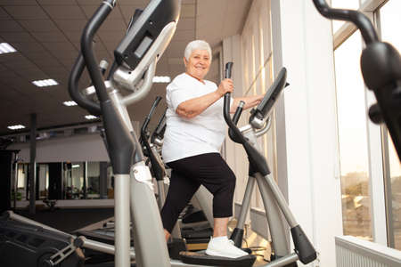 elderly retired woman with gray hair is engaged in sports on simulators in gym. Healthy lifestyle, senior concept, indoor. Rehabilitation, treatment after injuries, improvement. Individual lessons.の写真素材