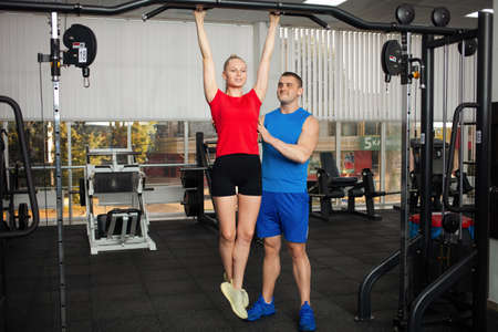 teenager performs an exercise on a simulator in gym. girl hangs on horizontal bar. coach shows right...