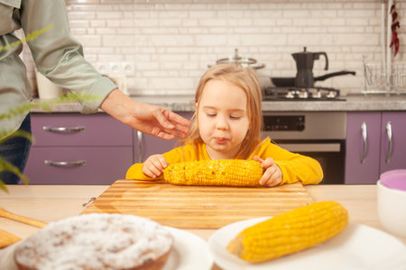 child eats sweet boiled corn in purple kitchen of house. Funny girl at table.の写真素材
