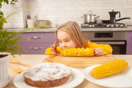child eats sweet boiled corn in purple kitchen of house. Funny girl at table. wrigglesの写真素材