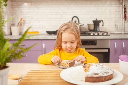 funny little girl eats plum pie with sugar, happy and satisfied, holding spoon on her own. purple kitchenの写真素材