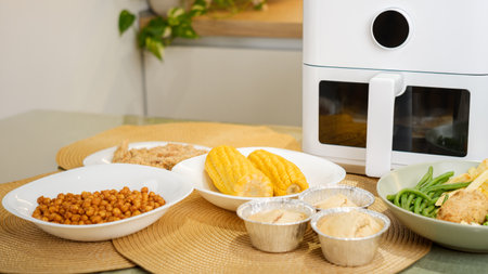 Air fryer sits on a kitchen counter surrounded by plates of cooked food, showcasing its versatility in preparing diverse mealsの写真素材