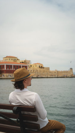 Tourist wearing hat sitting on bench admiring venetian fortress in chania, crete, greeceの写真素材