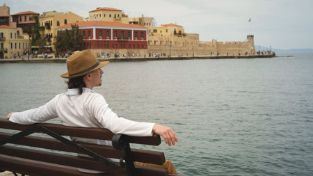Young man sitting on bench admiring the venetian harbor in chania, creteの写真素材