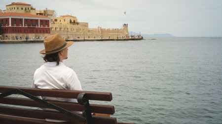 Young man sitting on a bench admiring the venetian fortress koules in creteの写真素材