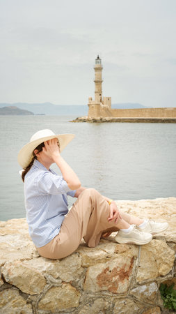 Woman wearing sun hat sitting on a stone wall enjoying the view of the venetian lighthouse in chania, creteの写真素材