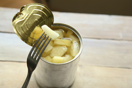 Opened tin can of canned pineapple pieces. Canned pineapple in can with fork on wooden table.の写真素材
