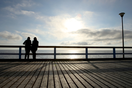 Silhouette of two girls or women on the bridge at sunset. Two friends standing on a wooden pier and looking at the sea.の写真素材