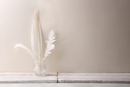 White quill feathers in glass bottle on white wood table. Feathers against white wall with empty place.の写真素材