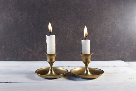 Burning two candles in vintage metal candlesticks on white wooden table against dark stone...