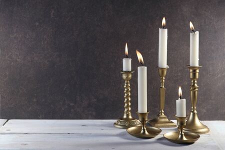 Burning candles in vintage metal candlesticks on white wooden table against dark stone background with copy space.
の写真素材
