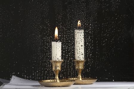  Burning two candles behind the glass with raindrops. Drops of water on the glass and candles in...